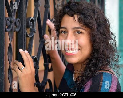 Belle positive émotive jeune mexicaine Latina hispanique femme avec de longues boucles noires maintient à un noir de sécurité de la grille de fenêtre en acier peint. Banque D'Images