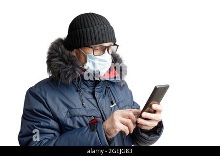 Homme en vêtements d'hiver, chapeau noir, lunettes et masque de protection tient un smartphone dans ses mains et regarde sur l'écran. Isolé sur blanc. Banque D'Images