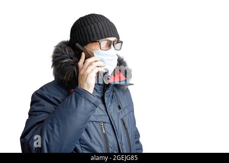 Homme en vêtements d'hiver, chapeau noir, lunettes et masque de protection parlant sur le smartphone. Isolé sur blanc. Copier l'espace. Banque D'Images