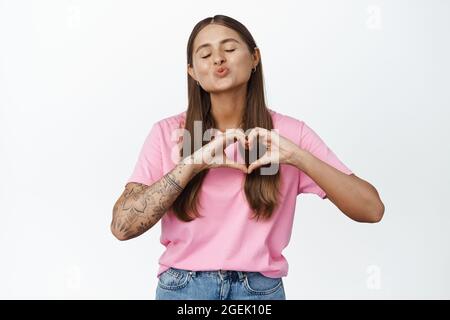 Jeune femme avec des tatouages pucker lèvres pour baiser, montre le coeur main signe et les yeux proches, debout sily contre fond blanc Banque D'Images