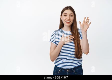 Jeune fille amicale souriant, levant une main, mettre le bras sur le coeur, se présenter, en disant bonjour, debout sur fond blanc Banque D'Images