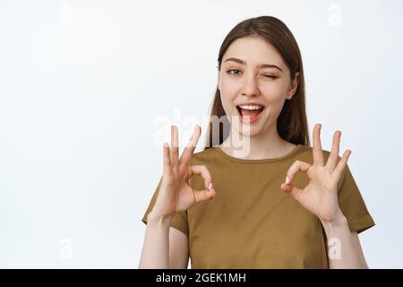 Image d'une fille souriant et montrant des signes corrects, en vous clin d'œil pour confirmer smth, tout sous contrôle, tout bon, en assurant la qualité, en se tenant dessus Banque D'Images