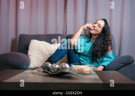 Vue horizontale d'une femme regardant un film à la maison, elle est assise sur son canapé très regardant la caméra et mange beaucoup de pop-corn Banque D'Images