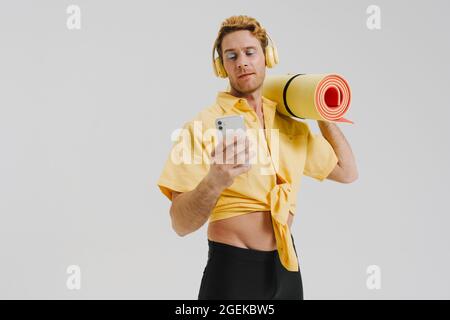 Jeune homme gay blanc portant un tapis de fitness écoutant de la musique avec un casque posé sur un fond de mur gris tenant un téléphone portable Banque D'Images