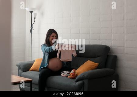 Femme enceinte Latina tenant et regardant les vêtements de son futur bébé. Banque D'Images