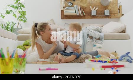 Une maman souriante et heureuse joue avec son bébé, avec des crayons et craies colorés, un jeu éducatif, assis à la maison entre des oreillers et des peluches d'animaux, Banque D'Images