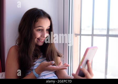 Jolie fille brune, assise près de la fenêtre ensoleillée de sa maison, sourit et gestuelle avec sa main, tout en effectuant un appel vidéo avec son smartphone. Banque D'Images