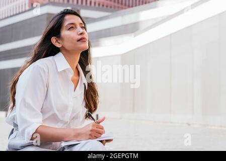 portrait horizontal d'une jeune femme indienne attrayante. Elle est assise près d'un immeuble de bureaux et regarde avec attention tout en tenant un ordinateur portable Banque D'Images