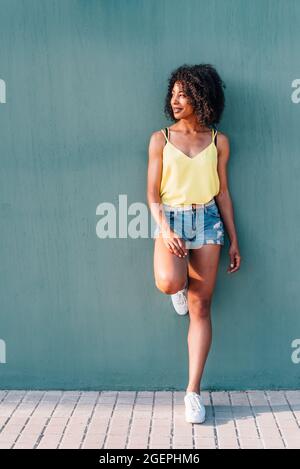 portrait vertical d'une femme africaine avec un fond de mur vert. Elle se penche sur le mur vert avec des vêtements d'été et souriant Banque D'Images