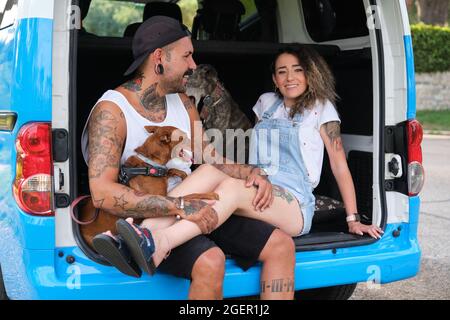 Jeune couple tatoué souriant et embrassant leurs chiens assis à l'arrière de la camionnette. Banque D'Images