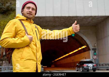 road trip, travel, gesture and people concept. jeune homme caucasien hitmarche et arrêt de voiture avec pouce vers le haut geste à la campagne, en manteau jaune Banque D'Images