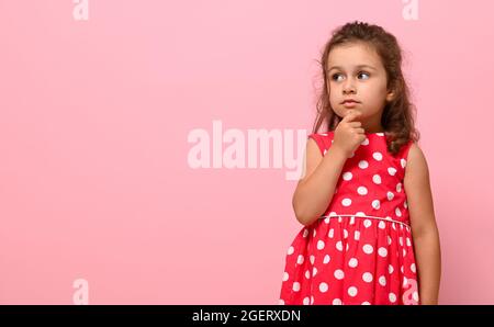 Une adorable petite fille d'âge préscolaire, de 3-4 ans, regarde soigneusement sur le côté, en tenant son doigt sur son menton, posant sur un backgroun rose Banque D'Images