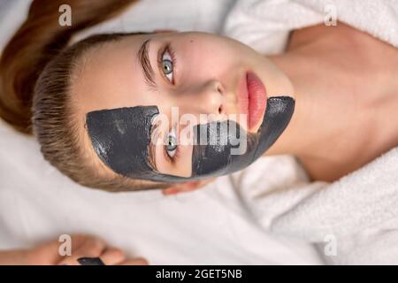 Top vue sur une jolie femme avec masque facial noir sur la moitié du visage, magnifique femme en peignoir blanc est couché sur le lit au spa, regardant la camisole Banque D'Images