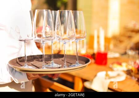 Photo rognée du serveur du service de restauration portant une chemise blanche portant un plateau rond marron avec un ensemble de verres à vin vides propres à la table des clients, o Banque D'Images