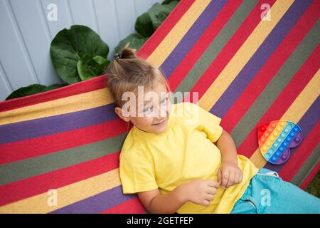 Garçon de 5 ans assis sur un hamac multicolore, joue avec un jouet Pop IT en forme de coeur. L'enfant est en vacances Banque D'Images