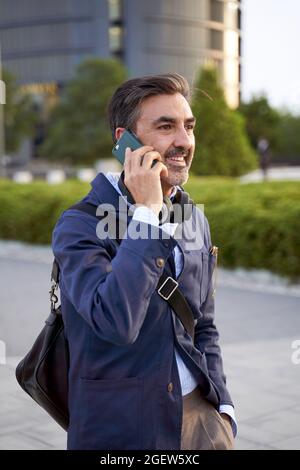 Homme mi-âge aux cheveux gris souriant heureux en utilisant un smartphone marchant dans la rue de la ville. Photo de haute qualité Banque D'Images