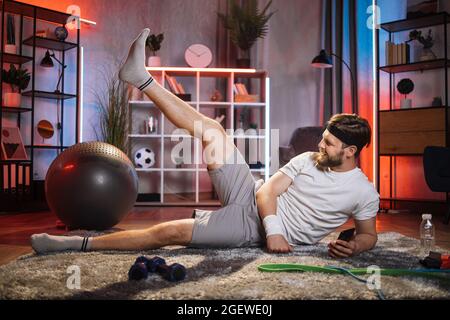 Jeune positif de bonne humeur sportif barbu allongé sur le sol et faisant des exercices de levage de jambe avec la jambe redressée pendant l'entraînement à la maison Banque D'Images