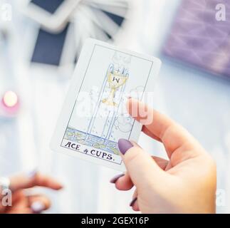 Main femelle avec clous violets tient la carte de tarot appelé As de tasses sur la surface blanche avec cahier et bougie. Vue de dessus. Minsk, Bélarus - 07.28.2021 Banque D'Images