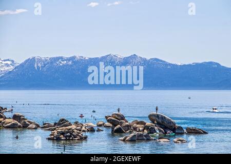 Touristes dans l'eau sur des paddleboards et escalade sur des rochers dans le lac Tahoe bleu avec des montagnes enneigées à distance Banque D'Images