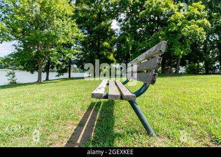 Un vieux banc de parc en bois et en métal se détériorant dans un champ d'herbe lors d'une journée ensoleillée d'été entourée de grands arbres. Banque D'Images