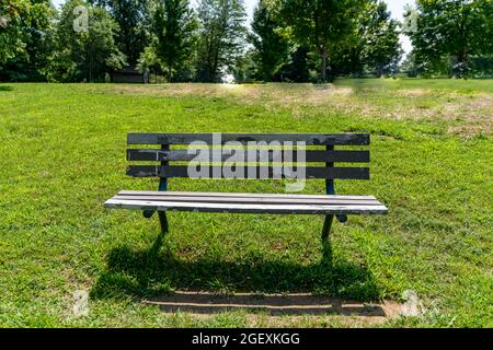 Un vieux banc de parc en bois et en métal se détériorant dans un champ d'herbe lors d'une journée ensoleillée d'été entourée de grands arbres. Banque D'Images