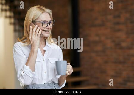 Femme d'âge moyen occupée avec des cheveux blonds et des lunettes dans une chemise blanche buvant du café ou du thé tout en parlant au téléphone, debout à la maison Banque D'Images