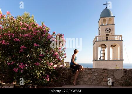 Femme assise près d'une oléande fleurie sur un petit mur, regardant le clocher de l'église et belle vue sur la mer et le village, Dhermi, Albanie Banque D'Images