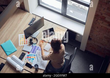Vue en grand angle de la créatrice féminine à l'aide de notes adhésives, travaillant sur un nouveau projet de design d'intérieur, assis dans un bureau moderne Banque D'Images