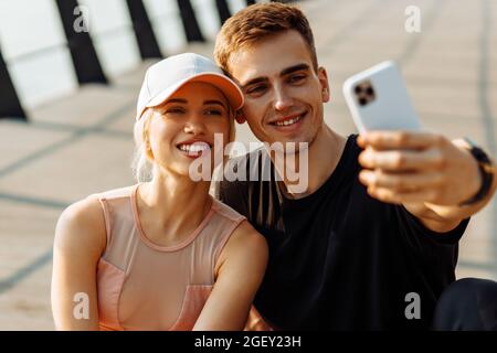 Les jeunes sportifs séduisants, homme et femme, se reposant après l'entraînement, prennent le selfie sur un téléphone portable, assis sur un tapis de fitness à l'extérieur sur la jetée Banque D'Images