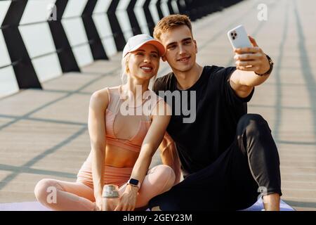 Les jeunes sportifs séduisants, homme et femme, se reposant après l'entraînement, prennent le selfie sur un téléphone portable, assis sur un tapis de fitness à l'extérieur sur la jetée Banque D'Images
