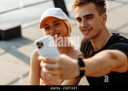 Les jeunes sportifs séduisants, homme et femme, se reposant après l'entraînement, prennent le selfie sur un téléphone portable, assis sur un tapis de fitness à l'extérieur sur la jetée Banque D'Images