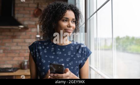 Bonne jeune femme afro-américaine pensive avec un smartphone qui regarde loin Banque D'Images