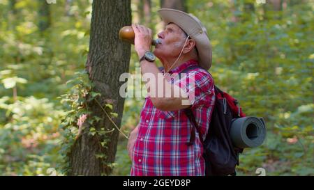 Fatigué assoiffé ancien randonneur caucasien grand-père relaxant, boire de l'eau dans la forêt après la marche Banque D'Images