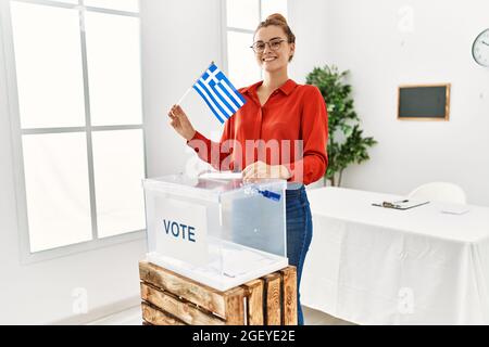 Jeune femme brune mettant une enveloppe dans une boîte de scrutin tenant le drapeau de la grèce à l'air positif et heureux debout et souriant avec un sourire confiant montrant Banque D'Images