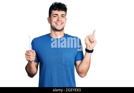 Jeune homme hispanique tenant une brosse à dents électrique souriant avec qui il se fait plaisir de pointer main et doigt sur le côté Banque D'Images