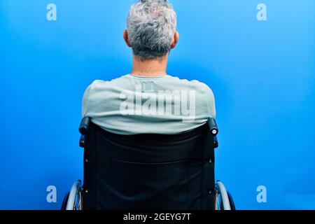 Beau homme d'âge moyen aux cheveux gris assis sur un fauteuil roulant debout en arrière en regardant loin avec des bras croisés Banque D'Images