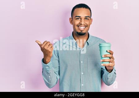 Un jeune afro-américain boit une tasse de café à emporter pointant le pouce vers le côté souriant heureux de bouche ouverte Banque D'Images