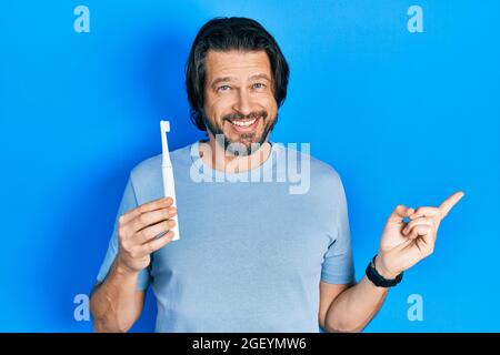 Homme de race blanche d'âge moyen tenant une brosse à dents électrique souriant heureux pointant avec la main et le doigt sur le côté Banque D'Images