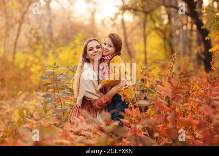 Photo de famille de la jeune belle mère hugs souriant petite fille crouching dans une grande herbe colorée dans la forêt d'automne, maman et enfant en chaud tricoté clo Banque D'Images