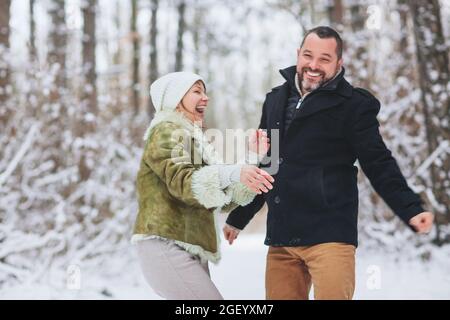 Charmant couple de familles d'âge moyen qui s'amusent en plein air en hiver, se promener dans la forêt enneigée avec le sourire, vêtu Banque D'Images