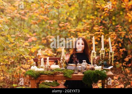 Petite fille adorable en costume de sorcière noire jouant dans la forêt d'automne, enfant s'amusant à table en bois avec mousse, citrouilles et diverses décorations, lo Banque D'Images