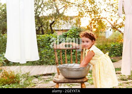 Mignonne petite fille en robe laver les vêtements blancs dans le bassin en métal dans l'arrière-cour, enfant aidant avec des corvées, pendant la lessive sur la corde à linge et la laissant t Banque D'Images