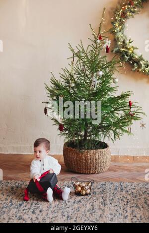 Adorable petit garçon tout-petit assis sur un tapis sous un arbre de noël décoré et déballant une boîte cadeau noire avec ruban rouge pendant la saison des fêtes d'hiver, W Banque D'Images