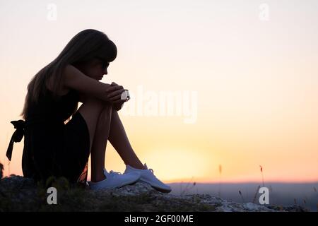 Jeune femme dépressive noire robe courte d'été assise sur une colline de montagne pensant à l'extérieur au coucher du soleil. Femme solitaire contemplant dans une soirée chaude Banque D'Images