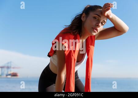 Gros plan d'une jeune femme de fitness attirante prenant une pause pendant le jogging, panant et essuyant la transpiration du front, regardant fatigué à l'appareil photo, debout Banque D'Images