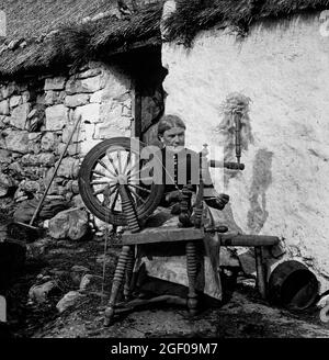 Scène du début du XXe siècle d'une femme qui tourne du lin à l'extérieur de son chalet de chaume en Irlande du Nord. Banque D'Images