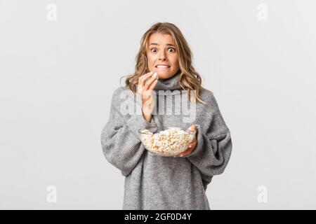 Portrait de fille blonde mangeant du pop-corn et regardant peur, regardant le film, debout sur fond blanc Banque D'Images