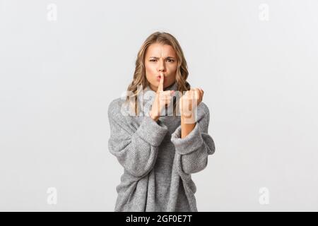 Gros plan d'une jeune fille blonde en colère et grognante dans un chandail gris, qui claque quelqu'un pour être bruyant, secouant la poing et le shushing, se tenant sur un fond blanc Banque D'Images