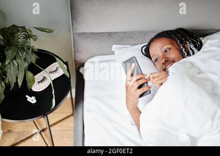 Portrait d'une jeune femme afro-américaine qui vérifie les messages de son smartphone après s'être réveillée le matin et avoir souriant, dans un espace de copie Banque D'Images