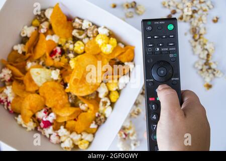 Vue de dessus d'une main tenant la télécommande du téléviseur contre le pop-corn frit et les frites sur un bol Banque D'Images